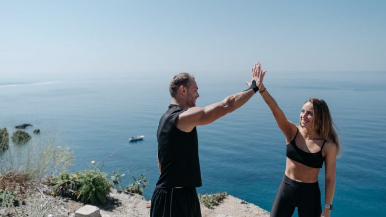 A happy couple in activewear high fives on a cliff by the ocean, enjoying a sunny day outdoors.